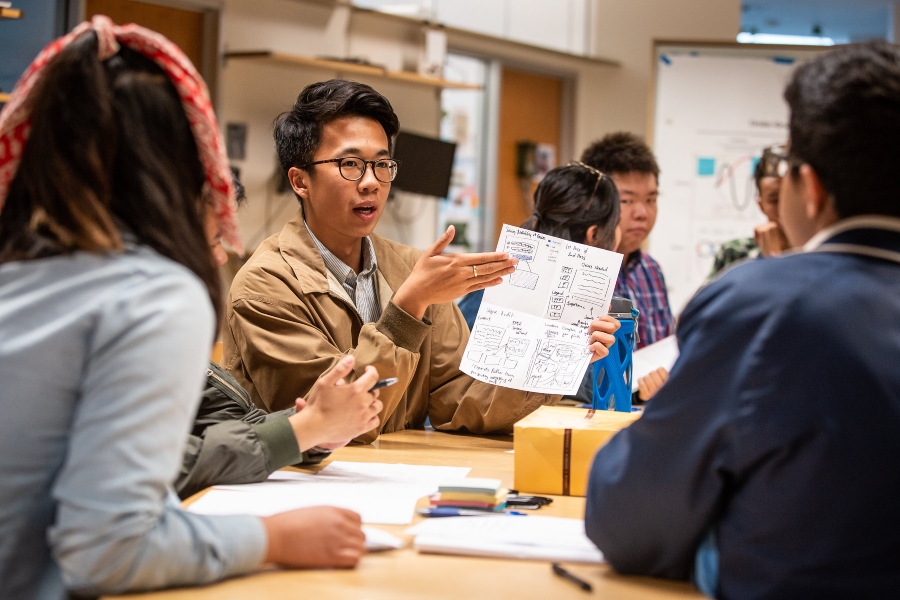 A student engaged in a group discussion holds up a paper displaying a wireframe for a new class registration interface.