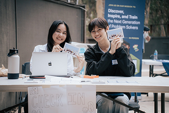 Two Design Lab student members sit behind a check-in table, smiling for a picture while holding up postcards