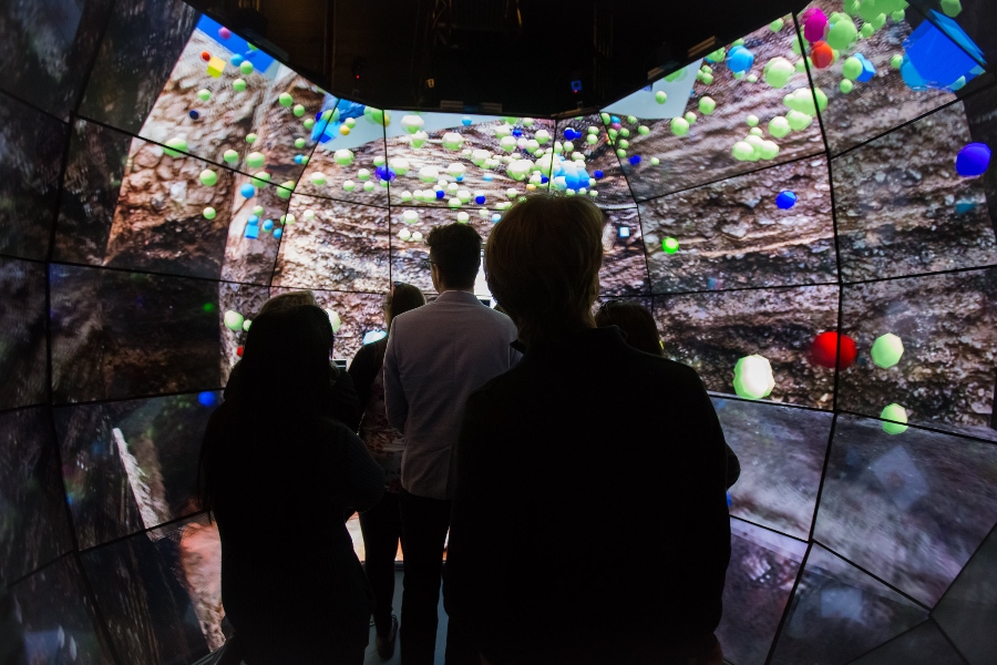 Participants at the Sonos visit at The Design Lab enter a room with floor-to-ceiling monitors, displaying an abstract image with multi-colored spheres scattered across the screen, creating an immersive experience.