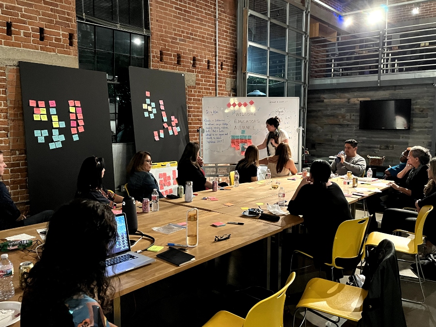 Participants at a UC San Diego design workshop sit around a table covered in post-it notes and markers, while one member adding a note to a whiteboard.